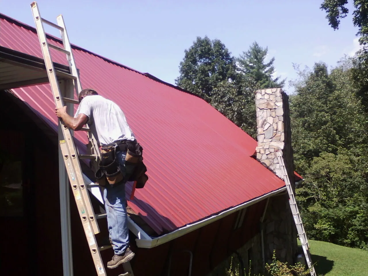 Roofer on a ladder beside a red metal roof and stone chimney — Soffit & Fascia Repair by Roofing Brothers in Loysville