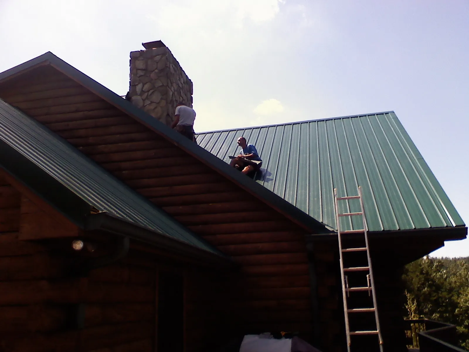 Roofers installing green metal panels beside a stone chimney on a log home in Loysville